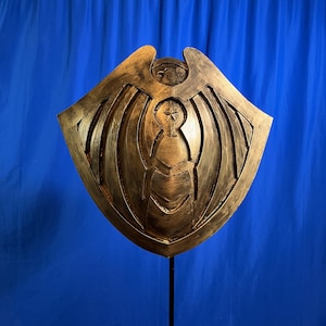 May include: A gold-colored shield with a detailed design featuring wings and a central figure. The shield is mounted on a black stand against a blue backdrop. The shield's surface has a textured appearance.
