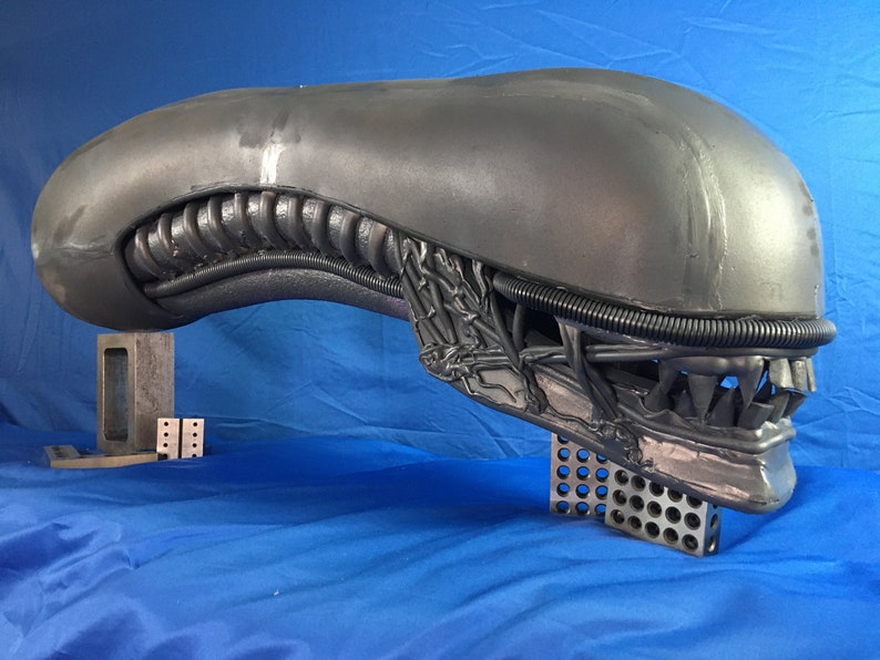 May include: A gray alien head sculpture with a detailed, textured surface. The head is mounted on a metal stand with a grid pattern.