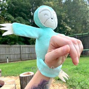May include: A light blue sloth hand puppet with white accents. The puppet has a smiling face and outstretched arms with white claws. The puppet is made of soft fabric and is designed to fit over the hand.