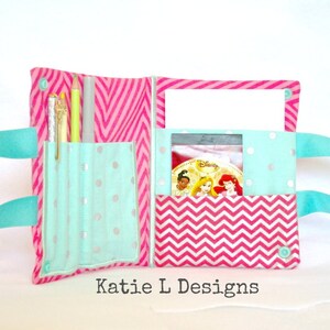May include: Open, handmade fabric pencil case with a teal handle. The case features a pink and white chevron pattern and teal fabric with silver polka dots. The case holds pens, pencils, and a Disney DVD. The text "Katie L Designs" is visible.