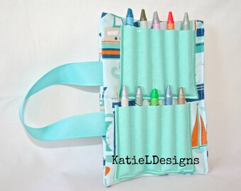 May include: A light blue fabric crayon holder with a sailboat pattern and a turquoise ribbon handle. The holder has eight slots for crayons and the text "KatieLDesigns" is printed on the front.