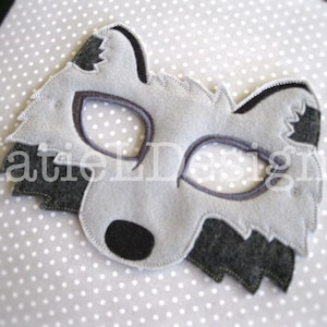 May include: A gray felt raccoon mask with dark gray accents and eye holes. The mask features a black nose and is set against a white polka dot background. The text "Katie LD Designs" is visible.