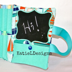 Könnte beinhalten: Eine Stofftafel mit dem Wort "Hi!" in weißer Kreide geschrieben. Die Tafel ist mit einem gewellten Rand umrahmt und an einem Stofforganizer mit Taschen und einem türkisfarbenen Bandgriff befestigt. Der Stoff hat ein nautisches Thema.