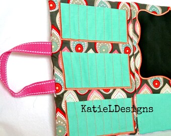 May include: A teal fabric organiser with three pockets and a chalkboard panel. The organiser has a pink handle and a paisley pattern with red, white, and grey accents. The text "KatieLDesigns" is printed on the bottom right corner.