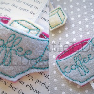 May include: A gray felt coffee cup bookmark with a teal embroidered coffee design and a pink felt interior. The bookmark is attached to a white ribbon.