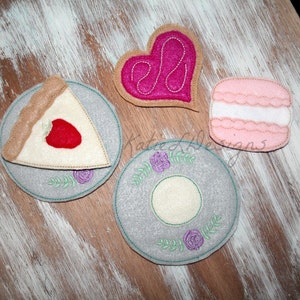 May include: Felt applique set with a heart, a slice of pie, a macaron, and two plates with floral designs. The heart is pink, the pie is beige with a red strawberry, the macaron is pink with white frosting, and the plates are gray with purple flowers.
