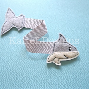 May include: A gray felt shark and tail attached to a gray and white chevron ribbon. The shark has a white belly and a small black eye. The ribbon is on a light blue background.