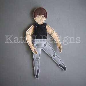 May include: A felt doll of a male ballet dancer with articulated limbs. The doll has brown hair, a black top, and gray pants. The doll is posed mid-dance with one leg extended. The doll's limbs are connected with small metal fasteners.