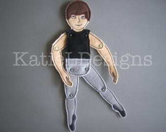 May include: A felt doll of a male ballet dancer with articulated limbs. The doll has brown hair, a black top, and gray pants. The doll is posed mid-dance with one leg extended. The doll's limbs are connected with small metal fasteners.