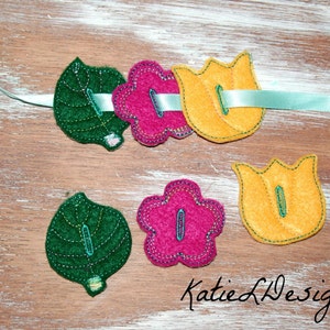 May include: A set of felt appliques featuring a green leaf, a pink flower, a yellow tulip, and a green leaf. The appliques are arranged in a row on a light wood surface. The appliques are perfect for adding a touch of whimsy to any craft project.