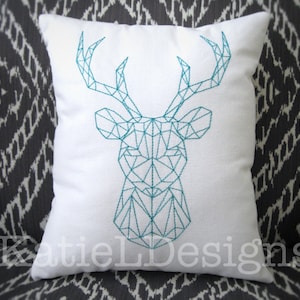 May include: A white throw pillow with a geometric teal deer design embroidered on the front. The pillow is sitting on a gray and white patterned couch.