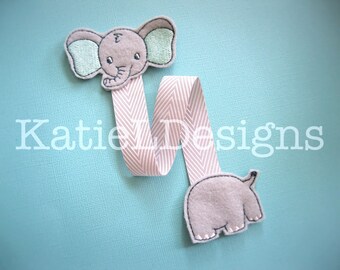 May include: A pink and white striped ribbon bookmark with an embroidered elephant on one end and a smaller embroidered elephant on the other end. The bookmark is on a light blue background.