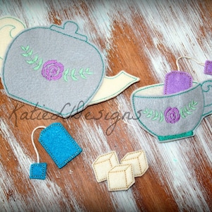 Può includere: Servizio da tè in feltro con teiera e tazza grigie, entrambe decorate con ricami floreali. Include bustine di tè blu e viola e zollette di zucchero beige. Il testo "Katie C Designs" è visibile.