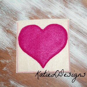 May include: A white felt coaster with a pink felt heart in the center. The coaster has a red stitched border and the text "Katie L Designs" is written in black cursive at the bottom.