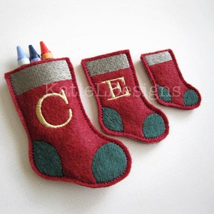 May include: Three red felt Christmas stockings with green trim and embroidered letters. The largest stocking has the letter "C" embroidered on it and has three crayons sticking out of the top. The middle stocking has the letter "E" embroidered on it. The smallest stocking has no letter embroidered on it.