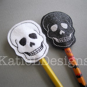 May include: Two Halloween-themed pencil toppers, one black and one white, both shaped like skulls with embroidered details. The pencils are yellow and orange with the text "Happy Halloween" on the orange pencil.