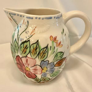 May include: A white ceramic pitcher with a curved handle and spout. The pitcher is decorated with hand-painted flowers in shades of pink, blue, and yellow, with green leaves. The rim has a blue dotted design. A decorative piece for the kitchen or dining room.