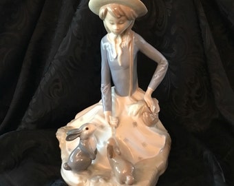 May include: A porcelain figurine of a girl wearing a straw hat and a blue dress, sitting with two rabbits. The girl is holding a basket in her hand.