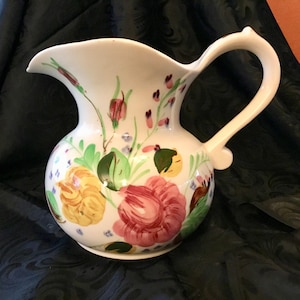 May include: A white ceramic pitcher with a floral design. The pitcher has a wide mouth and a handle. The flowers are painted in shades of pink, yellow, green, and purple.