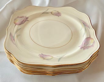 May include: A stack of square, off-white plates with a scalloped edge and gold trim. Each plate features a delicate floral design of purple tulips and green stems. The plates are stacked on top of each other.