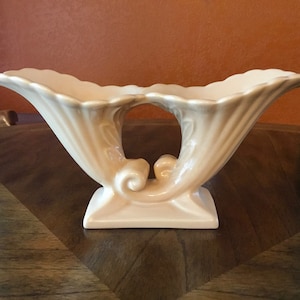 May include: A cream-colored ceramic vase with a unique, abstract design. The vase has two curved, trumpet-shaped openings and a decorative, scroll-like handle.