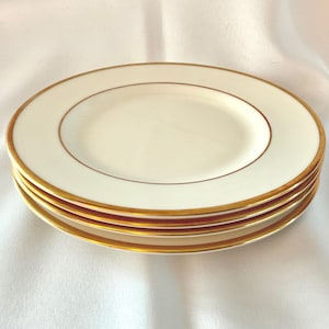 May include: A stack of ivory-colored plates with a gold rim and a thin gold inner line. The plates are stacked on top of each other, showcasing the elegant design. The plates are a classic addition to any dining set.