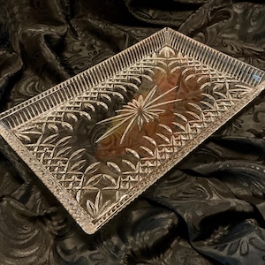 May include: A rectangular clear glass tray featuring an intricate pressed pattern of geometric shapes and leaf-like motifs, centered with a prominent starburst design and finished with a textured, serrated edge border.