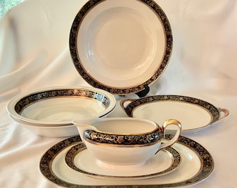 May include: A collection of white porcelain dinnerware with a black and gold floral design. The set includes a large serving bowl, a gravy boat with a saucer, and two oval serving platters. The plates have a gold rim.