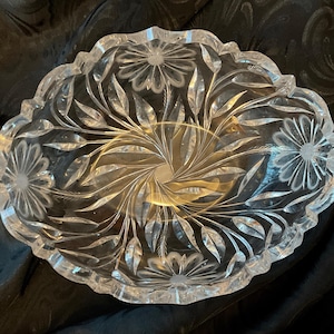 May include: Clear, oval-shaped crystal bowl with a scalloped edge. The bowl features intricate floral and leaf designs etched into the glass. The center has a spiral pattern. The bowl is approximately 10 inches in diameter.