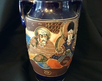 May include: A blue and white ceramic vase with a Japanese design featuring two figures. The vase has two handles and a rounded body.