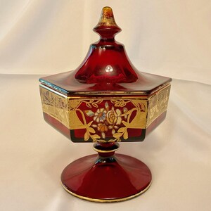 May include: A ruby red, lidded glass candy dish with a pedestal base. The hexagonal body is adorned with gold filigree and floral designs. The lid has a gold finial. The dish is an elegant decorative piece.