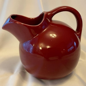 May include: A glossy, maroon-colored ceramic pitcher with a rounded body and a curved handle. The pitcher has a spout for pouring and is likely used for serving beverages. The design is simple and elegant, suitable for various kitchen styles.