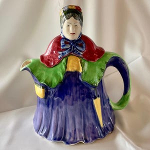 May include: A decorative ceramic teapot in the shape of a woman. The teapot features a red cape, green and blue accents, and a blue dress. The handle and spout are integrated into the design.