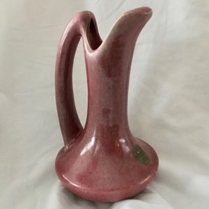 May include: A vintage, pink ceramic pitcher with a curved handle and a slender, elongated spout. The pitcher has a bulbous base and a smooth, glossy finish. A small, oval sticker is affixed to the side.