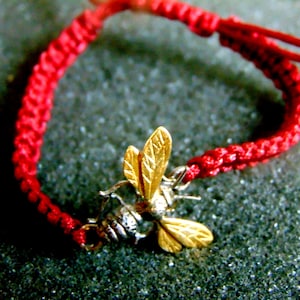 May include: A red woven bracelet featuring a gold-colored bee charm. The bee has detailed wings and a silver-colored body. The bracelet is made of a thick, braided red cord.