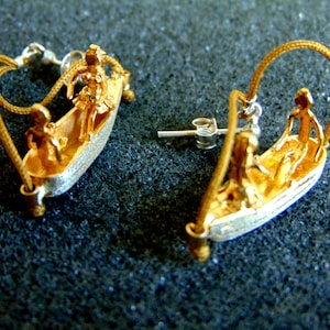 May include: A pair of gold earrings featuring a small boat with three figures inside. The boat is suspended from a gold wire loop. The earrings are made of metal and have a polished finish.