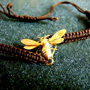 May include: A brown cord bracelet with a gold bee charm. The bee is detailed with wings and a body.