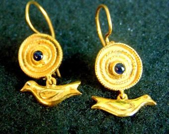 Peut inclure: Paire de boucles d'oreilles dorées. Chaque boucle d'oreille présente un motif circulaire avec une pierre bleue foncée au centre et un pendentif oiseau. Le fond est vert foncé.