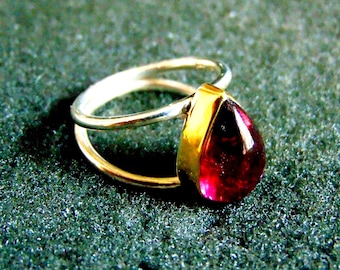 Könnte beinhalten: Ein silberner Ring mit einem goldenen Band und einem tropfenförmigen roten Edelstein.