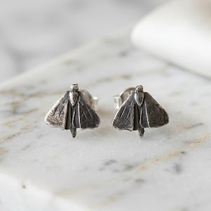 May include: A pair of silver stud earrings shaped like moths. Each earring features a detailed, textured wing design with a dark, oxidized finish that highlights the intricate lines and form of the insect, complete with a small post backing.