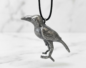 May include: A silver-toned metal pendant shaped like a standing crow or raven, featuring detailed wing and feather textures. The bird figure is suspended from a simple black cord necklace, creating a contrast between the metallic finish and the dark cord.