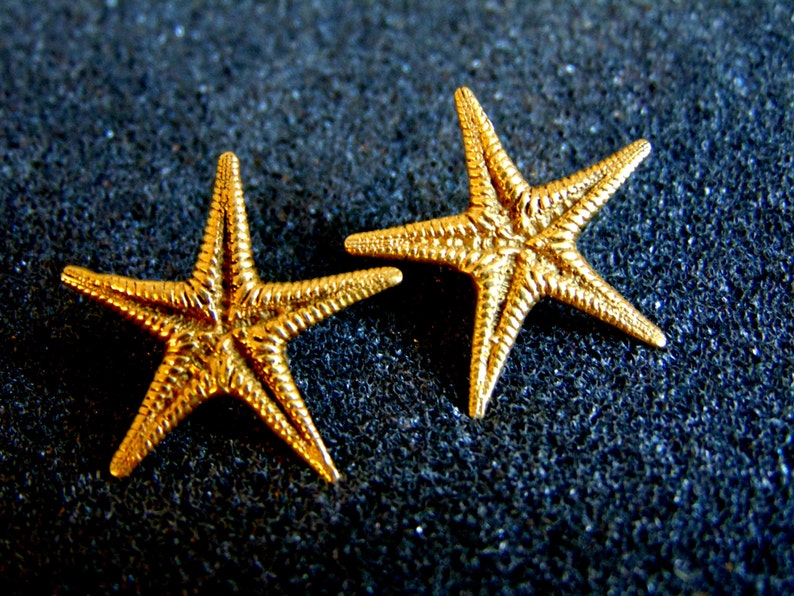 May include: Two gold-colored starfish earrings. The starfish are detailed with raised lines and have a slightly textured surface.