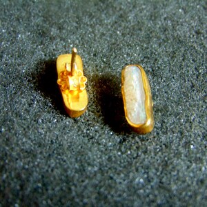 May include: A pair of gold-coloured rectangular stud earrings. One earring shows the post and backing. The other shows the face of the earring. The earrings have a light-coloured inlay, possibly a stone.