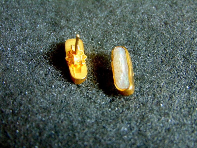 May include: A pair of gold-coloured rectangular stud earrings. One earring shows the post and backing. The other shows the face of the earring. The earrings have a light-coloured inlay, possibly a stone.