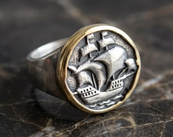 May include: A silver ring featuring a circular face with a raised relief of a sailing ship. The ship design is set against a dark, textured background and is framed by a smooth, polished gold-toned metal border that contrasts with the silver band.