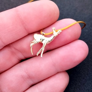 May include: A gold-colored pendant necklace featuring a deer charm. The deer is in a standing pose with its head raised. The necklace has a thin, yellow cord. The background is a dark color.