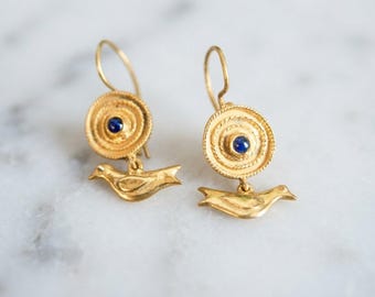 Peut inclure: Une paire de boucles d'oreilles pendantes dorées composées d'un disque circulaire avec une pierre bleue centrale et d'une breloque en forme d'oiseau. Les boucles sont suspendues à des crochets et présentent une finition métallique texturée.