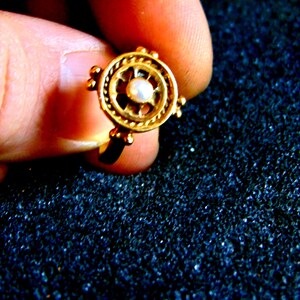 May include: A gold-colored ring with a circular design. The ring features a central pearl surrounded by an intricate pattern. Small, round accents are placed around the outer edge of the ring. The ring is held by a person's fingers.