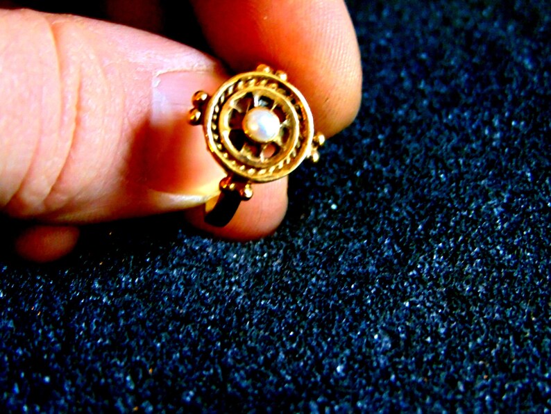 May include: A gold-colored ring with a circular design. The ring features a central pearl surrounded by an intricate pattern. Small, round accents are placed around the outer edge of the ring. The ring is held by a person's fingers.
