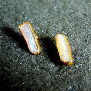 May include: A pair of gold-plated earrings, each with a rectangular design. They feature a natural, iridescent pearl at the centre. The pearls have an irregular shape. The earrings are displayed on a dark, textured surface.